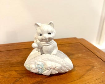 May include: A white ceramic cat figurine seated on a decorative pillow. The cat has a scarf and is holding a small object. The pillow has floral and ribbon details with light blue accents. The figurine is on a wooden surface.