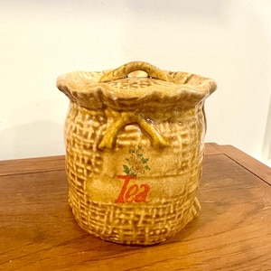 May include: A tan ceramic tea canister shaped like a woven bag with a lid and handle. The word "Tea" is printed on the front in red script, with a small floral design above it. The canister has a textured surface.