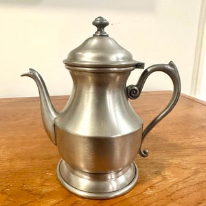 May include: A silver-toned metal teapot with a lid and a curved handle. The teapot has a spout and sits on a round base. The design is classic and elegant, suitable for serving tea.