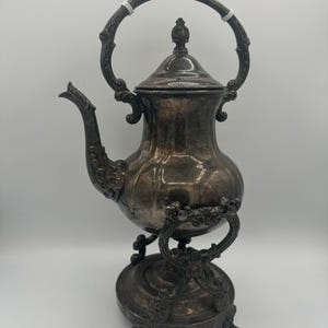 May include: Vintage silver-plated teapot with intricate detailing. The teapot has a curved spout, a domed lid with a finial, and a large handle. It rests on a decorative stand with three feet. The design is vintage and elegant, suitable for serving tea.