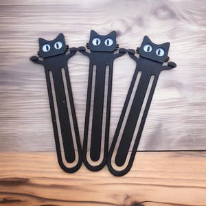 以下が含まれることがあります： 白い目をした黒い猫の形をしたブックマークが3つ。ブックマークは金属製で、シンプルなデザインです。
