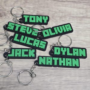 May include: Keychains with names in a pixelated font. The names "TONY", "STEVE", "OLIVIA", "LUCAS", "JACK", "DYLAN", and "NATHAN" are displayed in green on black rectangular keychains with silver keyrings.