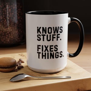 May include: A white ceramic mug with a black interior and handle, featuring the black text "KNOWS STUFF. FIXES THINGS." printed in a distressed, sans-serif font on the front. The mug is positioned next to a small spoon and a brown cookie.
