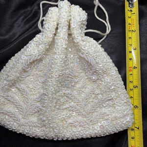 May include: Ivory beaded drawstring bag. The bag is covered in small, round sequins and seed beads, with a floral pattern. The bag is approximately 18 cm tall. The drawstring is made of a thin, off-white cord.