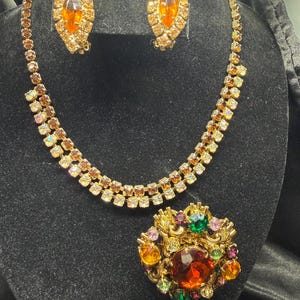 May include: A vintage jewellery set comprising a necklace, earrings, and a brooch. The necklace and earrings feature amber-coloured stones. The brooch has a large amber stone surrounded by smaller stones in various colours. The set is displayed on a black surface.