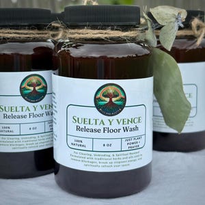 May include: Several glass bottles of "SUELTA Y VENCE Release Floor Wash" are displayed. Each bottle has a black cap, a label with a tree logo, and is wrapped with twine. The labels also include the text "100% NATURAL" and "8 OZ".