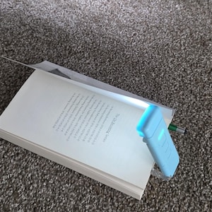 May include: A light blue LED book light clipped to a book. The book is open to a page with text. A green pen is also visible. The book and light are on a carpeted surface.