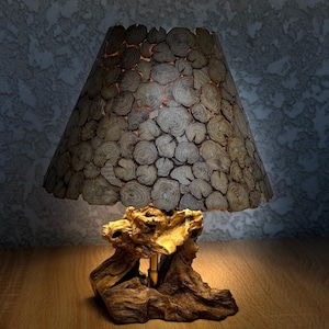 May include: A unique table lamp with a lampshade made of small, round wooden pieces. The base is crafted from a piece of natural wood, and the lamp emits a warm glow. The lamp is a decorative home decor item.