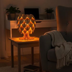 May include: A wooden table lamp with a leaf-shaped shade, emitting a warm light. The lamp is on a small wooden side table. The room features a gray armchair, a desk with a laptop, and potted plants, creating a comfortable setting.