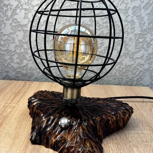 Puede incluir: Una lámpara de mesa única con una jaula de metal negro en forma de globo que rodea una bombilla de estilo vintage. La lámpara se asienta sobre una base de madera tallada de color marrón oscuro con un interruptor de encendido/apagado plateado.