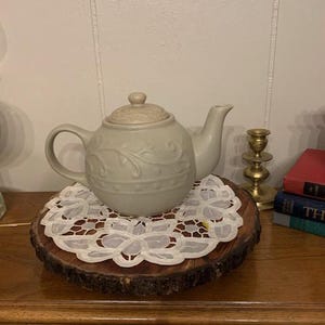 May include: A light green ceramic teapot with a floral design sits on a wooden slice with a white lace doily.