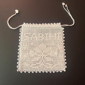 May include: A square, off-white crochet drawstring pouch featuring a delicate floral pattern in the center and the word "Sophie" woven into the top section. The edges are finished with a scalloped border and two thin matching crochet ties.