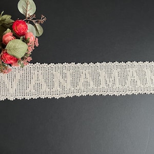 May include: A rectangular beige crochet runner featuring the word VANIAMAN in a repeating pattern, finished with a delicate scalloped edge. A small bouquet of artificial red roses and green eucalyptus leaves is positioned at the top left corner.