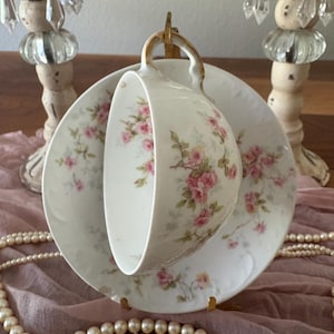May include: A white teacup and saucer set with a floral pattern of pink roses and green leaves. The cup is suspended above the saucer, showcasing the delicate design. The set has gold trim and is placed on a pink cloth.