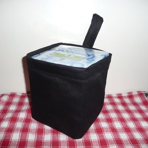 May include: A black, square tissue box cover with a handle. The cover is made of a solid black fabric and sits on a red and white checkered tablecloth. The top of the tissue box is visible, with the brand name "Scotts" printed on it.