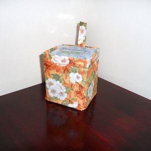 May include: A square tissue box cover with a floral pattern in orange, yellow, and green. The design features white flowers. A matching fabric handle is attached to the top. The box is sitting on a dark brown surface.