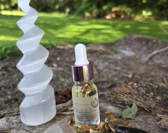 May include: A selenite spiral tower, a small essential oil bottle, a clear quartz crystal, and a black stone are arranged on a wooden surface. The oil bottle has a pink and gold cap and a label that reads "Back At Ya". The background is blurred.
