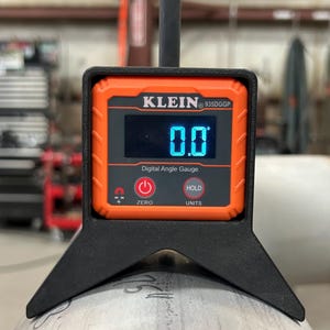 May include: A digital angle gauge by Klein Tools, model 935DGG, displays 0.0 degrees on a digital screen. The gauge is black and orange, with buttons for zero, hold, and units. It is resting on a metal pipe.