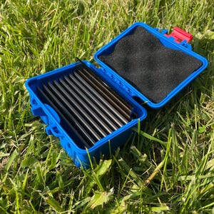 Pu&ograve; includere: Una scatola di plastica blu aperta, con un fermaglio rosso, contenente diversi oggetti rettangolari scuri. La scatola &egrave; foderata con schiuma nera e poggia su erba verde. La scatola misura circa 15 cm di lunghezza.