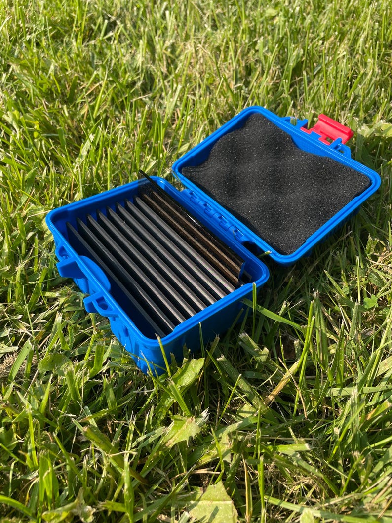 Pu&ograve; includere: Una scatola di plastica blu aperta, con un fermaglio rosso, contenente diversi oggetti rettangolari scuri. La scatola &egrave; foderata con schiuma nera e poggia su erba verde. La scatola misura circa 15 cm di lunghezza.