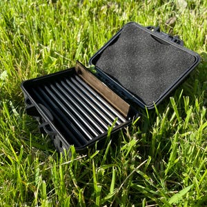 May include: An open black case with a foam interior and several silver metal objects inside, resting on a bed of green grass. The case has a rectangular shape with a hinged lid and a secure latch.
