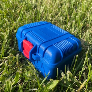 Pu&ograve; includere: Una scatola rettangolare blu con un fermaglio rosso &egrave; appoggiata sull'erba verde. La scatola presenta un design a coste sulla parte superiore e sui lati. Il fermaglio rosso &egrave; sul davanti.