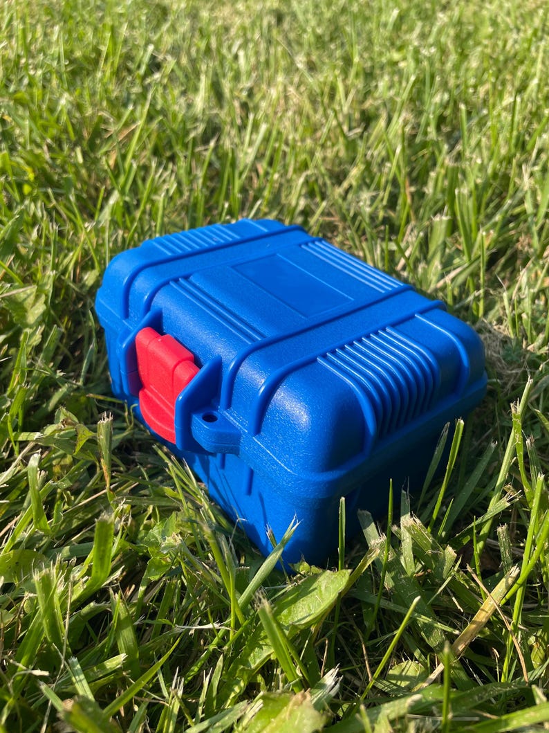 Pu&ograve; includere: Una scatola rettangolare blu con un fermaglio rosso &egrave; appoggiata sull'erba verde. La scatola presenta un design a coste sulla parte superiore e sui lati. Il fermaglio rosso &egrave; sul davanti.