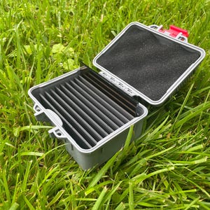 May include: An open, gray plastic fly fishing box with a black foam interior and gray dividers. The box has a red latch and is set on a bed of green grass.