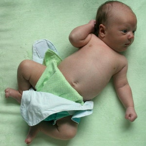 May include: A baby wearing a green and white patterned nappy cover lying on a light green blanket.