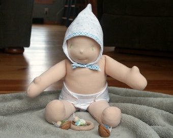 May include: A soft fabric doll with a light beige complexion, wearing a white bonnet with blue polka dots and a matching white diaper. The doll has simple embroidered facial features and is positioned near three small walnut shell toys.