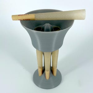 May include: A gray 3D-printed cone holder with three wooden legs and a round base. A pre-rolled cone is resting on top. The holder has a flared top and a central cone-shaped opening.