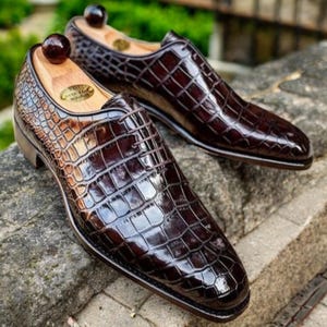May include: A pair of brown leather dress shoes with a crocodile skin pattern. The shoes are displayed on a stone surface, with wooden shoe trees inserted. The shoes have a glossy finish and are laced up.