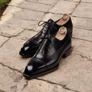 Op de afbeelding: Een paar gepolijste zwarte leren herenschoenen met een neus en decoratieve perforaties. De schoenen hebben veters en worden gepresenteerd met schoenspanners. Op een stenen oppervlak.