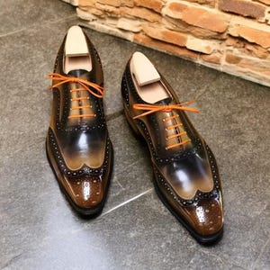 Może przedstawiać: Para brązowych i czarnych skórzanych butów wizytowych ze sznurowadłami w kolorze pomarańczowym. Buty mają design z ozdobnym skrzydłem i spiczasty nosek.