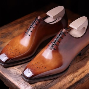 Può includere: Un paio di scarpe eleganti in pelle marrone con finitura lucida. Le scarpe hanno la punta quadrata, perforazioni decorative e lacci neri. Sono inserite forme di legno all'interno. Le scarpe sono esposte su una superficie di legno.