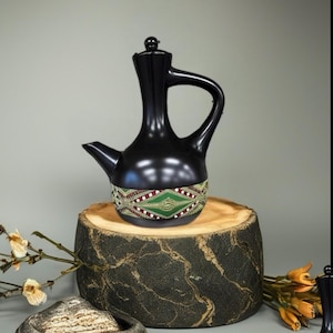 May include: A black ceramic coffee pot with a decorative band featuring green and red geometric patterns. The pot has a long spout and a curved handle. It sits on a wooden stump with a natural edge.