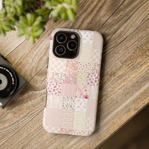 Könnte beinhalten: Eine Handyhülle mit Patchwork-Design in Rosa-, Creme- und Blumenmustern. Die Hülle hat einen Kameraausschnitt und liegt auf einer Holzoberfläche. Die Hülle ist ein schützendes Zubehör für ein Smartphone.