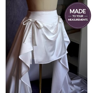 Könnte beinhalten: Weißer Satinrock mit hoher Taille und drapiertem Vorderteil. Der Rock hat Schleifendetails an der Taille und ein fließendes, asymmetrisches Design. Der Text "MADE - TO YOUR - MEASUREMENTS" befindet sich in einem lila Kreis.