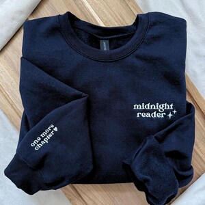 以下が含まれることがあります： 紺色のスウェットシャツ。前面に「midnight reader」という文字と2つの星、左袖に「one more chapter」という文字とハートマークがプリントされています。