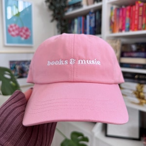 May include: A pink baseball cap with white embroidered text that reads "books & music".