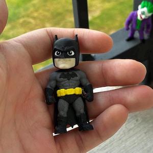 May include: A small Batman figurine featuring a black cowl, a grey textured suit with a black bat emblem on the chest, black gloves and boots, and a bright yellow utility belt. The character has a neutral facial expression with white eyes.
