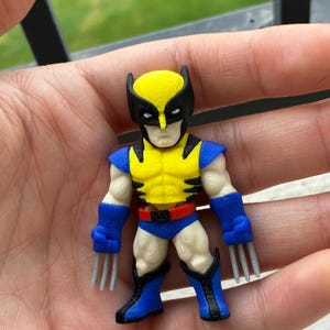 May include: A small, three-dimensional printed figurine of a comic book character featuring a yellow and black mask, a yellow chest piece with black stripes, blue shoulder pads, blue gloves with three silver claws on each hand, and blue boots.