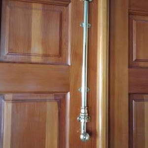 May include: A polished brass door bolt, vertically mounted on a wooden door. The bolt has a long, slender rod with decorative end pieces and mounting plates. The door has a natural wood grain finish.