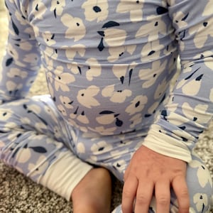Op de afbeelding: Lichtblauwe bloemenpyjamaset. De top met lange mouwen en de broek hebben een wit bloemenpatroon met marineblauwe accenten. De manchetten en de halslijn zijn wit. Het kind zit op een tapijt.