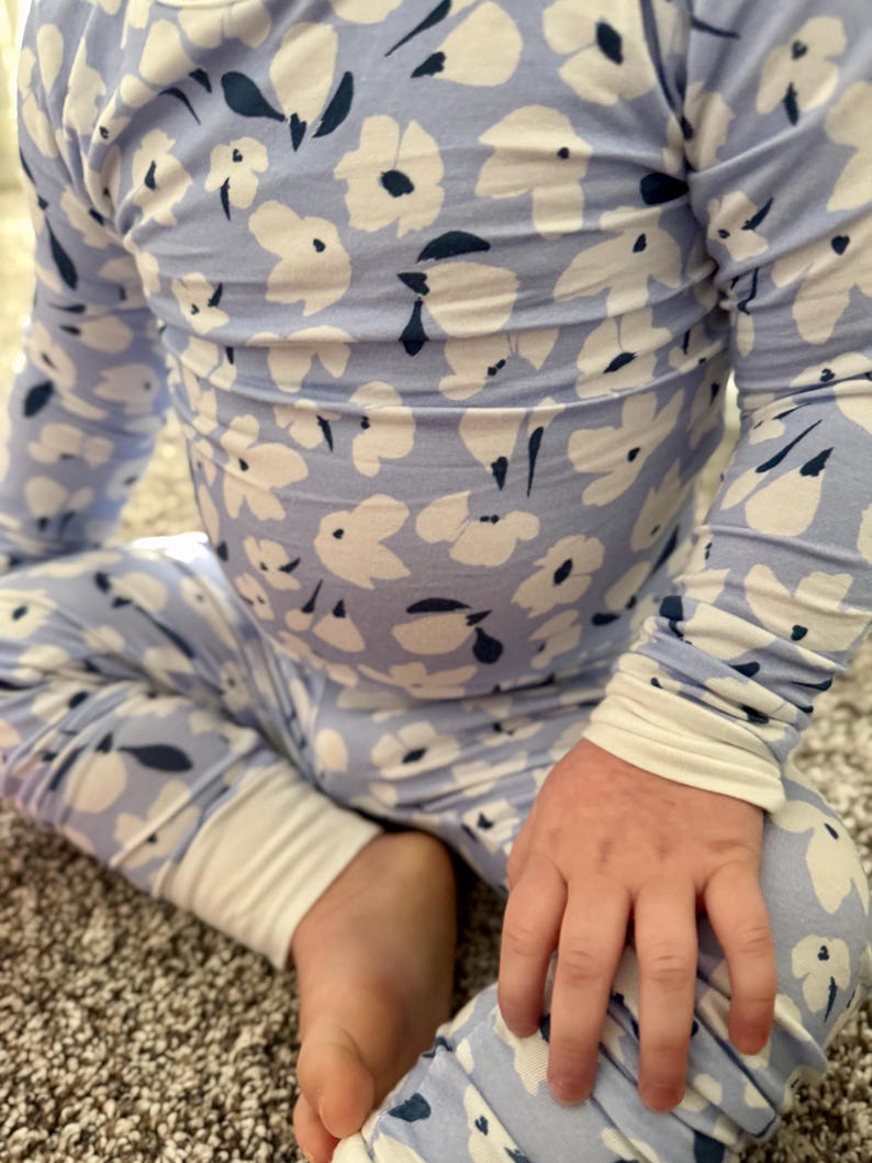 Op de afbeelding: Lichtblauwe bloemenpyjamaset. De top met lange mouwen en de broek hebben een wit bloemenpatroon met marineblauwe accenten. De manchetten en de halslijn zijn wit. Het kind zit op een tapijt.