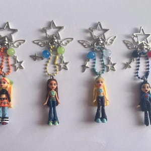 May include: Four doll keyrings with star and wing details. Each keyring features a miniature doll with different hair and clothing styles. The keyrings have colourful bead and star details, with a silver-tone star and wing design at the top.