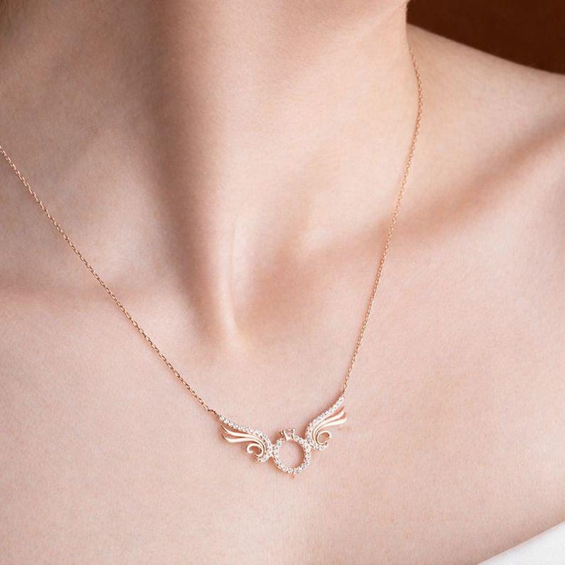 May include: A rose gold necklace featuring a pendant with a ring and wing design, adorned with small, sparkling stones. The necklace is displayed on a person's neck, showcasing its delicate chain and intricate details.