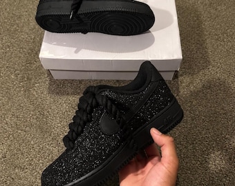 May include: A pair of black low-top sneakers featuring a glittery, textured upper, thick rope-style laces, and a solid black rubber sole. One sneaker is held in the foreground, while the other rests on a white cardboard box in the background.