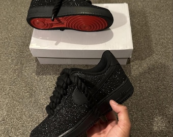 May include: A pair of black glitter sneakers featuring thick rope-style laces and a solid black midsole. One sneaker is positioned on a white box, revealing a bright red outsole, while the other is held up to show the side profile.