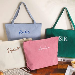 May include: Four personalized tote bags in various colors: light blue, green, pink, and off-white. Each bag features a name or initials embroidered in a contrasting color. The bags have long handles for carrying and are displayed on a wooden surface.